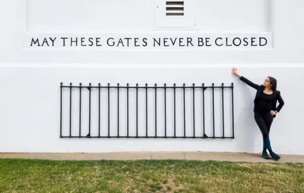 Wall with iron fence with inscribed "MAY THESE GATES NEVER BE CLOSED". A white woman in black gestures at the words.